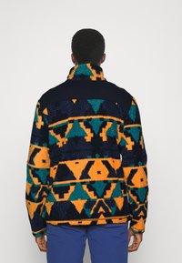 Veste en polaire avec des motifs géométriques multicolores en orange, turquoise et marine. Col haut et tissu texturé avec une coupe décontractée.