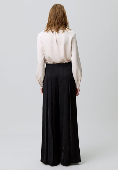 Jupe noire longue avec des plis. Tissu satiné brillant, taille haute et design fluide. Blouse légère de couleur claire avec des détails froncés au dos.