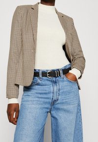 Brązowo-beżowa marynarka w kratę typu houndstooth o dopasowanym kroju, zestawiona z kremowym golfem, wysokim stanem niebieskich dżinsów i czarnym paskiem.