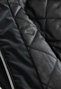 Giacca nera trapuntata in tessuto con una finitura lucida, caratterizzata da motivi a diamante, foderata di scuro e rifinita con zip argentata.