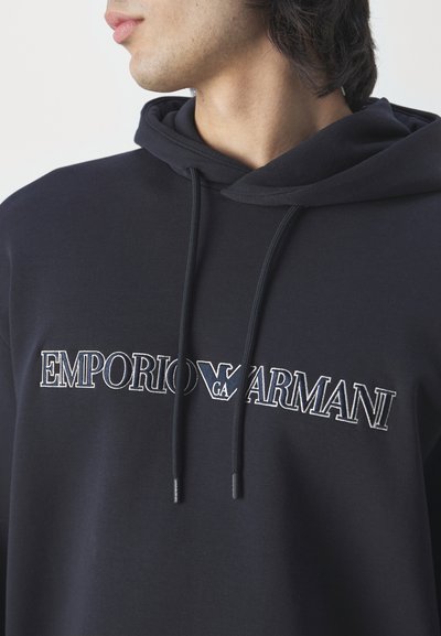 Felpa con cappuccio nera caratterizzata dal logo "EMPORIO ARMANI" ricamato in bianco e grigio chiaro. Il tessuto sembra morbido e presenta una vestibilità comoda.