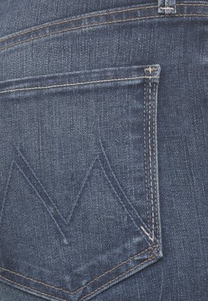 Primo piano della tasca posteriore di jeans di denim blu con cuciture visibili e motivo "M" ricamato sul tessuto.