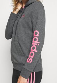 Sweat à capuche gris avec logo "adidas" rose sur la manche. Comprend une poche avant, une capuche avec cordon de serrage et un tissu doux et texturé.