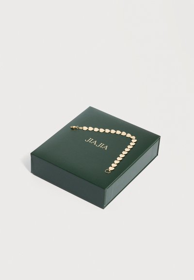 Χρυσό βραχιόλι με αλυσίδα σε σχήμα καρδιάς, εμφανιζόμενο σε σκούρο πράσινο κουτί κοσμημάτων με χρυσό ανάγλυφο όνομα της μάρκας, σε λευκό φόντο.