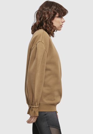 Kvinde med bølget brunt hår iført en løs, langærmet brun sweatshirt og sorte bukser, stående i profil mod en ensfarvet baggrund.