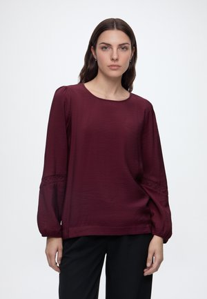 Femme aux longs cheveux foncés porte un chemisier bordeaux à manches longues bouffantes et un pantalon noir, debout devant un fond clair uni.