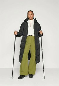 Schwarze Pufferjacke mit Kapuze, weiße Reißverschlussjacke und olivgrüne weitgeschnittene Hose. Accessoires sind Trekkingstöcke und schwarze Schuhe.
