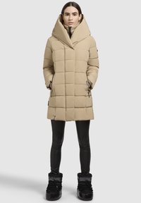 Beige pufferjakke med en fremtrædende hætte, der har et quiltet design, lynlåslommer og er parret med sorte leggings og støvler.