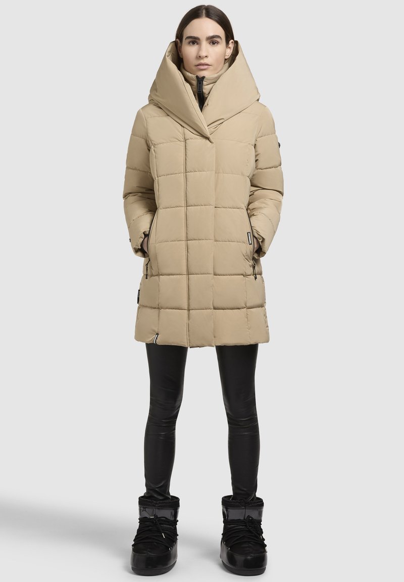 Beige Daunenmantel mit einer auffälligen Kapuze, ausgestattet mit einem gesteppten Design, Reißverschlusstaschen und kombiniert mit schwarzen Leggings und Stiefeln.