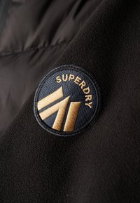 Chaqueta de forro polar negra con un parche bordado en forma redonda. El parche es de color azul marino con detalles dorados y contiene la palabra "SUPERDRY" y formas geométricas.