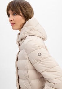 Beige Daunenjacke mit Kapuze, einem gesteppten Muster, subtiler Schimmer und einem dekorativen runden silbernen Akzent am Arm.