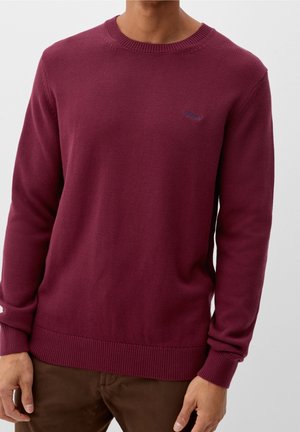 Mann trägt einen langärmeligen, bordeauxfarbenen Pullover mit Rundhalsausschnitt, kombiniert mit brauner Hose, stehend vor einem schlichten hellen Hintergrund.