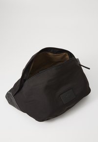 Borsa in tessuto nero con una superficie liscia, chiusura con zip e un interno marrone. Presenta una patch con logo e una base strutturata in materiale a contrasto.
