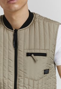 Beige quiltet vest med svart ribbestrikket krage, frontglidelås og lomme med glidelås på venstre bryst. Har et minimalistisk merke nederst.