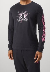 Schwarzes Langarm-T-Shirt mit pinkem Aufdruck einer Person, die in der Mitte springt, und einem komplizierten pinken Design am rechten Ärmel.