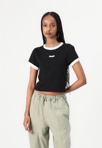 Svart cropped t-skjorte med hvit ribbestrikket hals og ermekanter, med en liten logo på brystet. Bært med lysegrønne bukser.