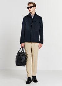 Giacca navy con colletto alto e due tasche sul petto, abbinata a pantaloni beige, scarpe nere e una borsa nera con texture.