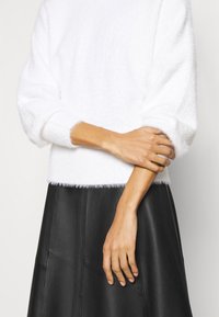 Weißer flauschiger Pullover mit weicher Textur und weiten Ärmeln, kombiniert mit einem schwarzen Lederminirock, der geometrische Einsätze für Kontraste aufweist.