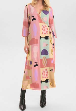 Femme portant une robe portefeuille aux couleurs pastel avec des motifs abstraits de vase et de tasse, associée à des bottines noires, se tenant devant un fond uni.