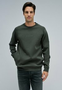 Felpa verde scuro con dettagli a coste sulle spalle, dotata di collo rotondo e logo ricamato "Salsa Jeans" sul davanti. Abbinata a jeans grigi.