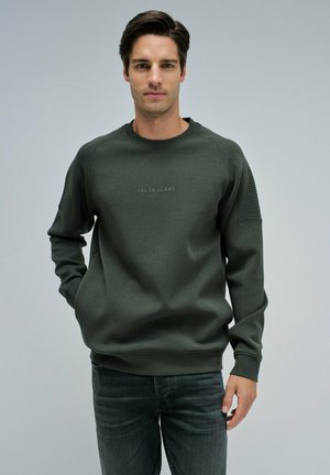Sudadera verde oscura con detalles acanalados en los hombros, que cuenta con cuello redondo y un logo bordado de "Salsa Jeans" en la parte frontal. Combinada con unos pantalones vaqueros grises.