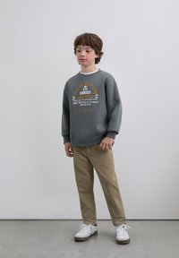 Gray sweatshirt featuring a front graphic with text and a skull, paired with tan pants and white sneakers. Smooth fabric with a relaxed fit.