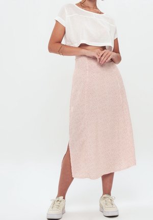 Vrouw draagt een wit cropped t-shirt, roze midi rok met kleine stippen en een zijsplit, en witte sneakers, staand met ineengestrengelde handen.