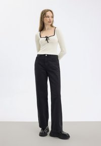 Top lungo beige chiaro a maniche lunghe con scollatura quadrata e bordi neri, abbinato a pantaloni neri a gamba larga e calzature nere chunky con borchie.
