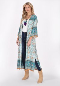 Kimono lungo floreale in turchese e crema, con maniche larghe, bordi arancioni e spacchi laterali, indossato sopra una maglietta bianca e jeans scuri.