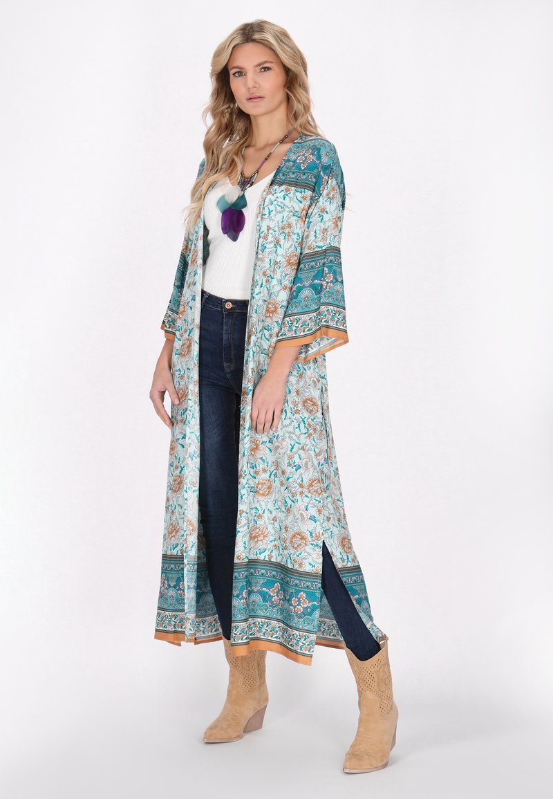 Kimono lungo floreale in turchese e crema, con maniche larghe, bordi arancioni e spacchi laterali, indossato sopra una maglietta bianca e jeans scuri.