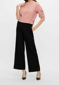 Femme portant un pull en maille torsadée rose, un pantalon noir large avec revers, et des talons pointus noirs, posant avec une main sur la hanche.