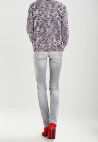 Osoba nosící vícebarevný pletený svetr, světlé šedé úzké džíny a červené vysoké podpatky, stojící zády k divákovi.