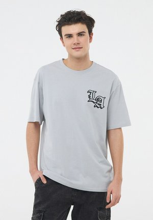 T-shirt grigio a maniche corte con grafica nera "LA" sul lato sinistro del petto, realizzato in tessuto morbido e con una vestibilità comoda.