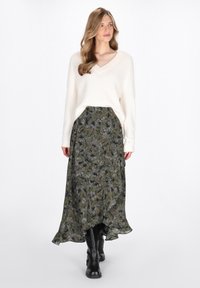 Donna con capelli lunghi che indossa un maglione bianco lavorato a maglia, una gonna midi a fiori verde decorata e stivali neri fino al ginocchio, che cammina in avanti su uno sfondo bianco.