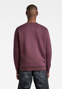 Sudadera marrón con cuello redondo, puños acanalados y banda de cintura. Presenta un parche de tela en la parte posterior del cuello y una etiqueta con el logo en el costado.
