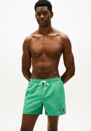 Skjorteløs mand iført lysegrønne badeshorts med hvid snor, stående med hænderne bag ryggen mod en ensfarvet hvid baggrund.