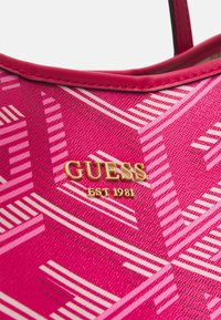 Roze handtas met een diagonaal gestreept patroon, voorzien van een gouden metalen "GUESS EST 1981" logo-plaat op textuurmateriaal.