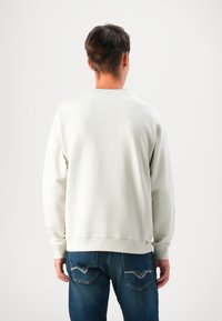 Man wearing plain white long-sleeve sweatshirt and blue jeans facing away against a plain white background.