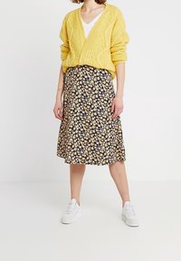 Un pull en maille jaune porté sur une chemise blanche à col en V, associé à une jupe noire ornée de petites fleurs jaunes, et des baskets blanches.