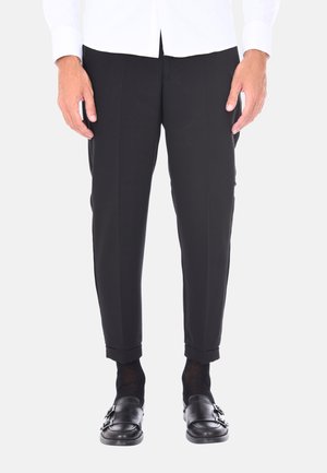 Pantaloni neri su misura con una texture liscia, caratterizzati da una vestibilità slim e orli risvoltati. Indossati con scarpe e calzini neri.