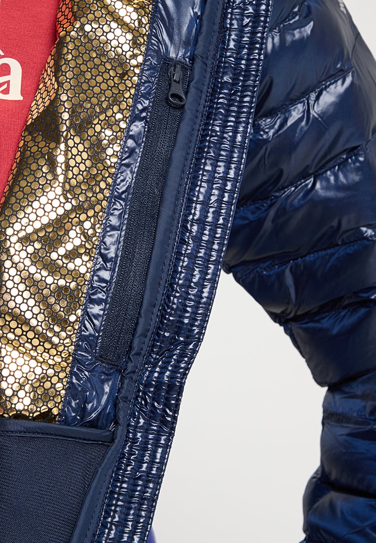 Veste matelassée bleu marine avec une doublure intérieure brillante en motif de nid d'abeille doré et une poche zippée intérieure à côté d'un tissu rouge.