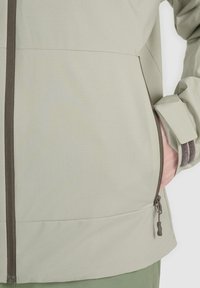 Hand partially inside side zip pocket of light beige jacket with dark brown zipper and cuff details.