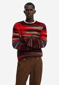 Czerwony sweter w paski w wielu kolorach z okrągłym dekoltem i długimi rękawami, zestawiony z brązowymi spodniami plisowanymi.