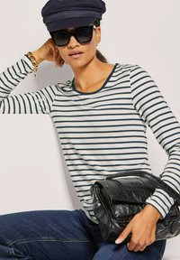 Gestreiftes Langarm-Shirt in Creme und Marineblau, runder Ausschnitt, kombiniert mit einer schwarzen Lederhandtasche und einer Marineblauen Kappe. Das Model trägt Sonnenbrillen.