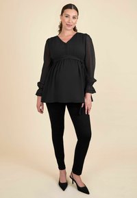 Čierny materinský top s textúrovanou tkaninou, výstrihom do V, detailom gombíka a dlhými priesvitnými rukávmi; skombinovaný s čiernymi úzkymi nohavicami a špicatými podpätkami.