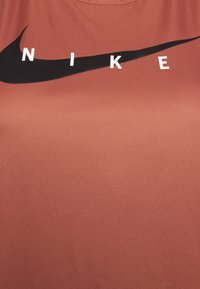 Nike T-shirt met korte mouwen, in terracottastof. Heeft een opvallende zwarte swoosh en witte letters die "NIKE" spellen. Gladde textuur.