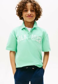 Polo shirt verde menta con maniche corte, caratterizzato da scritte e dettagli bianchi. Indossato con pantaloni blu scuro, che mostrano un look casual.
