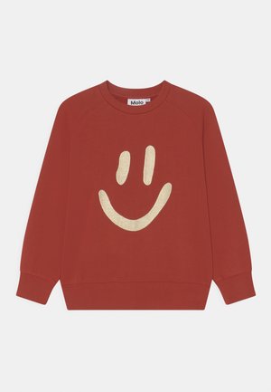 Sweatshirt rouge avec un col rond et des manches raglan. Présente un grand design d'un visage souriant doré sur le devant. Fabriqué en coton doux.