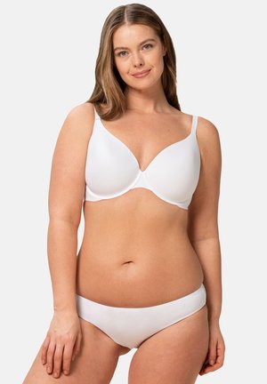 Donna sorridente con capelli lunghi che indossa un reggiseno bianco e biancheria intima bianca, in piedi contro uno sfondo chiaro e uniforme.