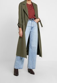 Oliivinvihreä trench-manteli, jossa on vyötärölle sidottu vyö, yhdistettynä marjapuuron väriseen toppiin ja vaaleansinisille leveälahkeisille farkuille. Tummat korkokengät viimeistelevät asun.